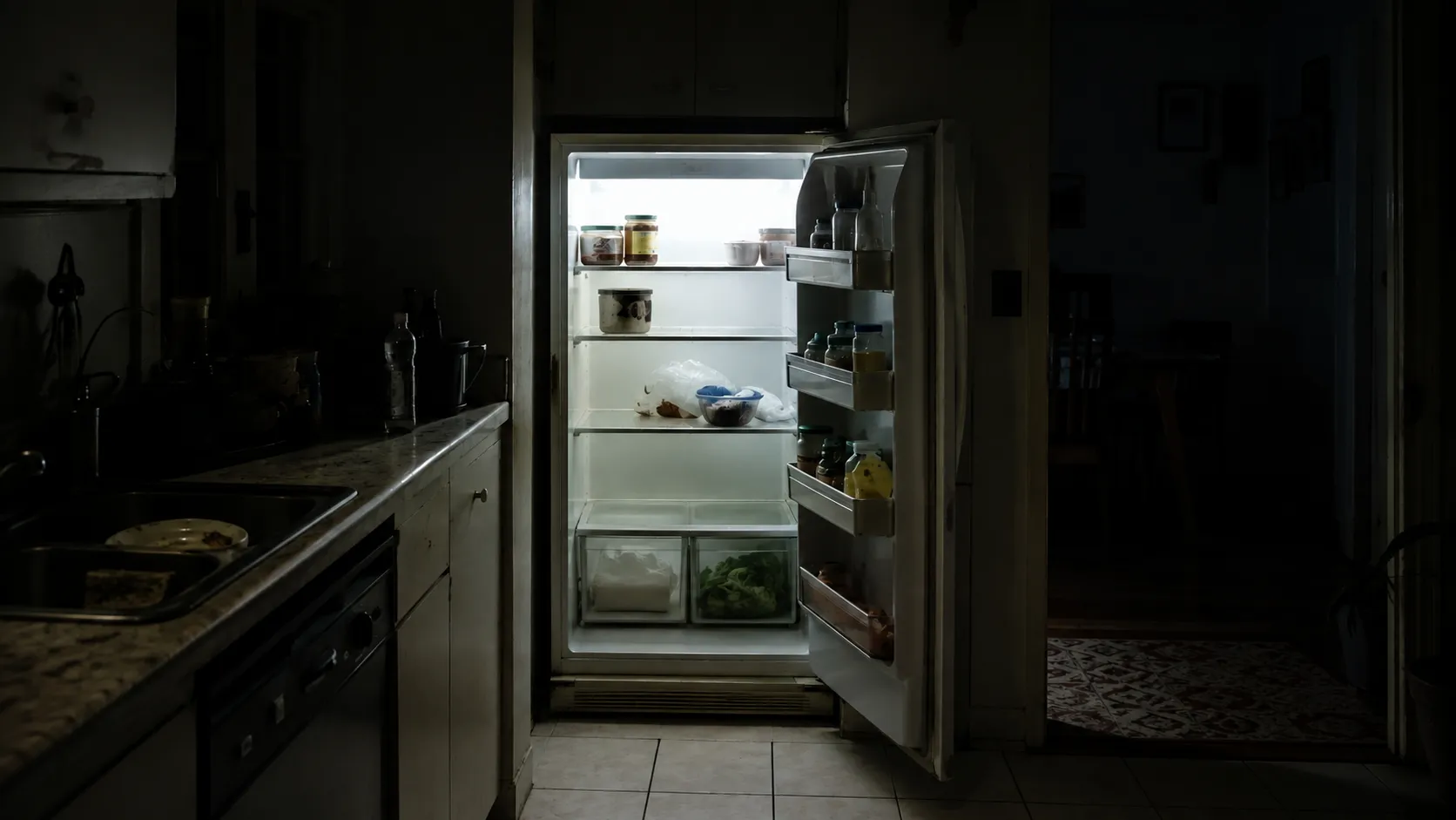 An open refrigerator at night in a dark kitchen, its shelves nearly empty